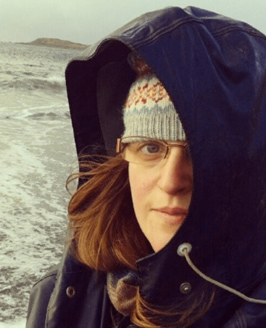 Photo of a white woman with glasses standing in front of a stormy sea with her face partly obscured by the hood of a jacket